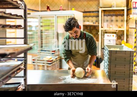 Boulangerie hispanique femelle pétrissant la pâte tout en se tenant debout au comptoir de métal dans la cuisine de boulangerie Banque D'Images