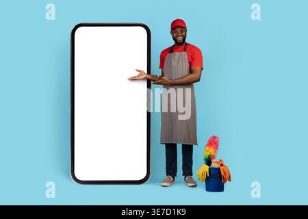Capuchon de port de nettoyant masculin noir et tablier pointant les mains sur le grand smartphone debout près du panier d'outils de nettoyage sur fond bleu dans le studio Banque D'Images