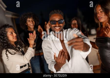 Homme noir portant des lunettes de soleil et chemise blanche dansant avec des amis sur le toit la nuit Banque D'Images