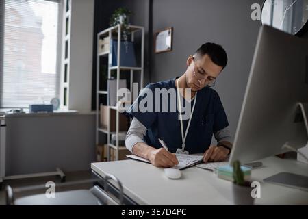 Médecin masculin mixte dans les gommages portant des lunettes remplissant la forme médicale sur le presse-papiers tout en étant assis à la table avec l'ordinateur en clinique Banque D'Images