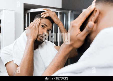 Inquiet homme noir avec serviette de bain sur les épaules touchant et vérifiant son cuir chevelu dans le miroir debout dans la salle de bain à la maison Banque D'Images