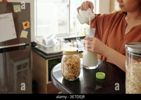 Image recadrée d'une femme asiatique versant du lait de la bouteille au pichet en verre debout dans la cuisine à la maison Banque D'Images