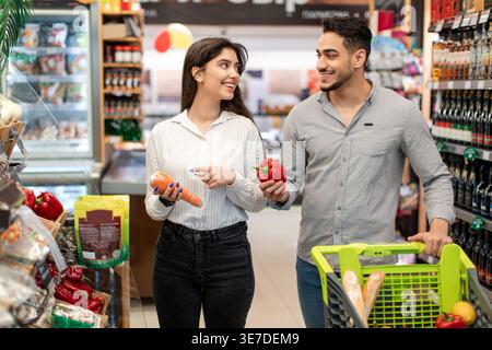 Couple souriant du moyen-Orient se regardant les uns les autres tenant des légumes tout en se tenant debout avec chariot dans le supermarché Banque D'Images