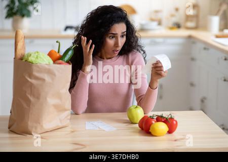 Femme choquée du moyen-Orient regardant le projet de loi gestuelle de la main tout en étant assise à la table près du sac en papier avec des produits d'épicerie dans la cuisine à la maison Banque D'Images