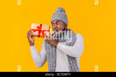 Homme noir étonné portant bonnet tricoté et écharpe élevant le cadeau de Noël à côté de son oreille debout sur fond jaune dans le studio Banque D'Images