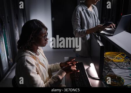 Ingénieur INFORMATIQUE féminin noir portant des lunettes de vue travaillant sur ordinateur assis à table près d'une collègue féminine caucasienne à l'aide d'un ordinateur portable tout en se tenant debout dans Ser... Banque D'Images