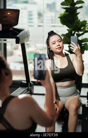 Femme asiatique prenant selfie avec smartphone assis devant le miroir dans le gymnase Banque D'Images