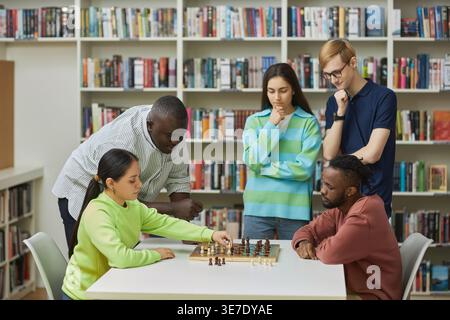 Femme hispanique jouant aux échecs avec un ami mâle noir assis à table près de collègues dans la bibliothèque du collège Banque D'Images