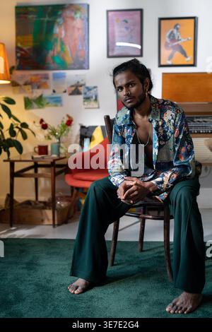 Artiste masculin indien sérieux assis pieds nus sur une chaise contre ses diverses peintures sur le mur dans le home studio Banque D'Images