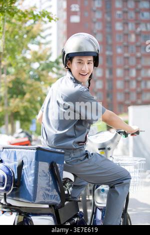 Homme chinois souriant portant un casque regardant par-dessus l'épaule tout en roulant sur scooter à moteur électrique avec sac de livraison sur la rue de la ville pendant la journée Banque D'Images