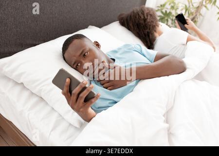 Homme noir utilisant un smartphone tout en étant allongé sur le lit contre sa petite amie dans la chambre à coucher à la maison Banque D'Images