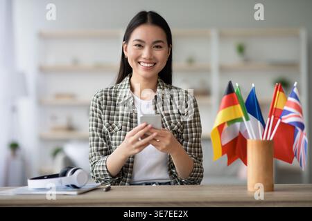 Femme asiatique souriante à l'aide d'un smartphone assis à table avec des écouteurs sans fil et des drapeaux internationaux à la maison Banque D'Images