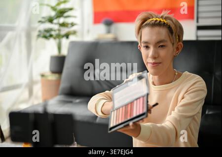 Homme gay asiatique montrant sa palette de fards à paupières sur la caméra tout en enregistrant le tutoriel de maquillage assis sur le sol près du canapé à la maison Banque D'Images