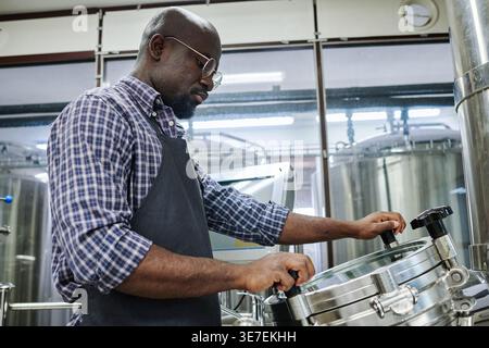 Homme chauve noir portant tablier opérant des machines debout dans l'usine Banque D'Images