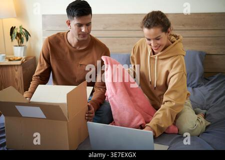 Femme de race mixte souriante utilisant un ordinateur portable et tenant une couverture assise près de petit ami asiatique sur le lit à la maison Banque D'Images