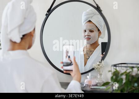 Femme noire avec masque facial portant un peignoir et les cheveux enveloppés dans une serviette prenant selfie à l'aide d'un smartphone regardant dans le miroir à la maison Banque D'Images