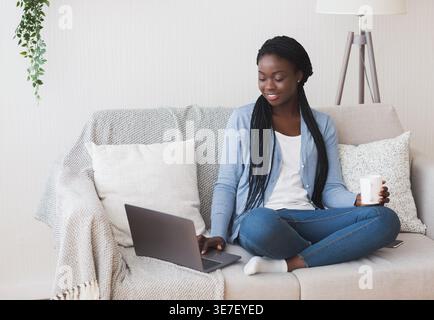Femme noire travaillant sur un ordinateur portable et tenant une tasse à café assise les jambes croisées sur le canapé travaillant à distance dans le salon à la maison Banque D'Images