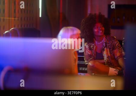 Souriante productrices de musique hispanique écoutant des chansons tout en étant assis ensemble dans un studio d'enregistrement Banque D'Images