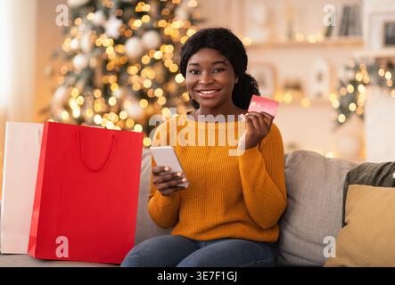 Femme noire tenant une carte de crédit et un smartphone assis sur un canapé avec des sacs à provisions à la maison Banque D'Images