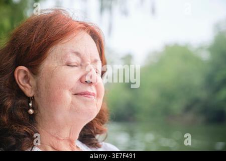 Femme âgée hispanique avec les yeux fermés debout près du lac pendant la journée Banque D'Images