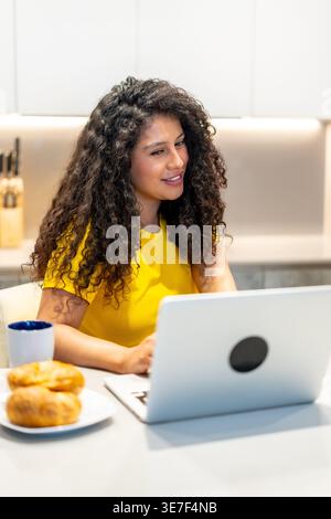 Femme hispanique avec les cheveux bouclés assister à la réunion en ligne à l'aide d'un ordinateur portable tout en prenant le petit déjeuner au comptoir de cuisine à la maison Banque D'Images