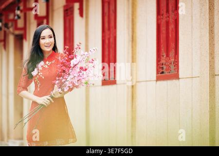 Femme asiatique souriante dans des vêtements traditionnels tenant des branches de pêche florissantes debout à l'extérieur Banque D'Images