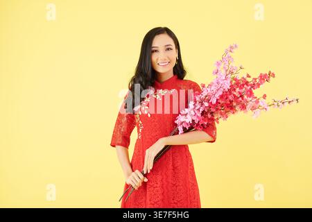 Femme asiatique souriante dans des vêtements traditionnels tenant des branches de pêche florissantes debout sur fond jaune dans le studio Banque D'Images