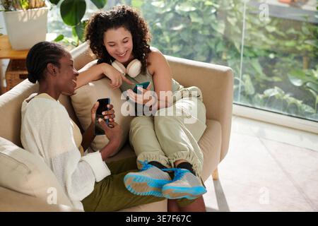 Femme mixte avec les jambes sur les genoux de colocataire femelle noire regardant la vidéo drôle sur les smartphones assis sur le canapé dans le salon pendant la journée Banque D'Images