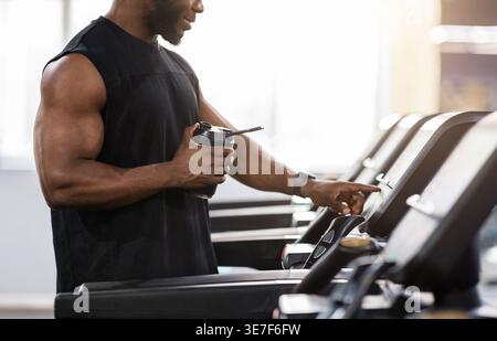 Image recadrée d'un homme noir tenant une bouteille d'eau sur un tapis roulant tout en se tenant debout dans la salle de gym pendant l'entraînement Banque D'Images