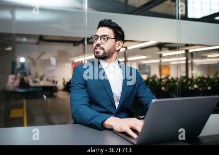 Homme du moyen-Orient avec barbe et lunettes regardant loin à l'aide d'un ordinateur portable assis au bureau dans le bureau Banque D'Images