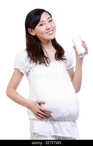 Femme enceinte chinoise souriante tenant le verre de lait debout sur fond blanc dans le studio Banque D'Images