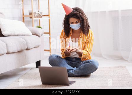 Contrarié femme noire portant chapeau de fête et masque médical tenant cupcake avec bougie et ayant appel vidéo sur ordinateur portable tout en étant assis sur la moquette du sol à la maison Banque D'Images