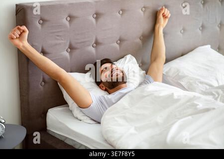 Homme arabe souriant avec la barbe portant une chemise blanche étirant les bras avec les yeux fermés tout en étant allongé sur le lit dans la chambre à coucher à la maison Banque D'Images