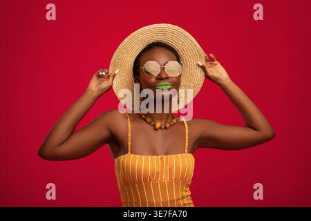 Femme noire avec les cheveux courts portant chapeau de paille et lunettes de soleil debout sur fond rouge dans le studio Banque D'Images