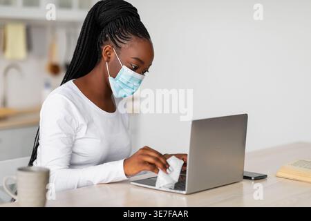 Femme noire avec des tresses portant un masque médical nettoyant son ordinateur portable tout en étant assise à table dans la cuisine à la maison Banque D'Images