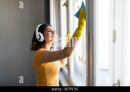 Adolescente hispanique portant des écouteurs et des gants en caoutchouc debout et utilisant un chiffon dans le nettoyage de la fenêtre à la maison Banque D'Images
