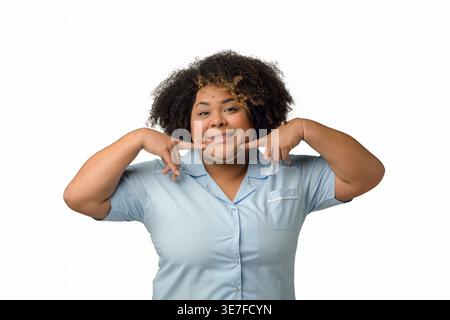 Femme hispanique de grande taille souriante avec afro bouclé portant un uniforme bleu montrant un sourire de doigt debout sur fond blanc dans le studio Banque D'Images