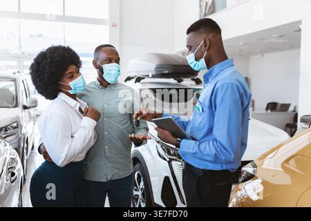 Couple noir dans les masques d'achat de nouvelle voiture et de prendre la clé du vendeur tenant la tablette tout en se tenant debout ensemble dans la salle d'exposition pendant la pandémie Banque D'Images