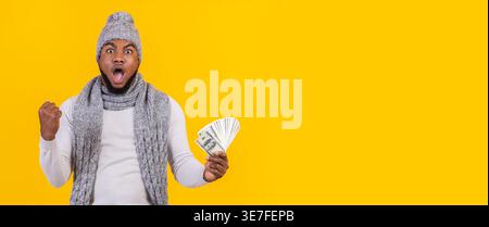 Homme noir choqué portant un bonnet tricoté et une écharpe levant le poing serré et tenant de l'argent célébrant tout en se tenant debout sur fond jaune en ... Banque D'Images