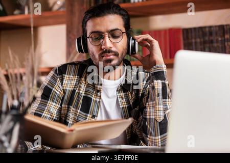 Étudiant indien portant des lunettes touchant des écouteurs lisant un livre assis à table dans la bibliothèque Banque D'Images