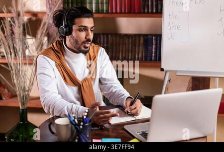 Enseignant indien dans des écouteurs écrivant des notes tout en enseignant en ligne à l'aide d'un ordinateur portable assis à la table dans la bibliothèque Banque D'Images
