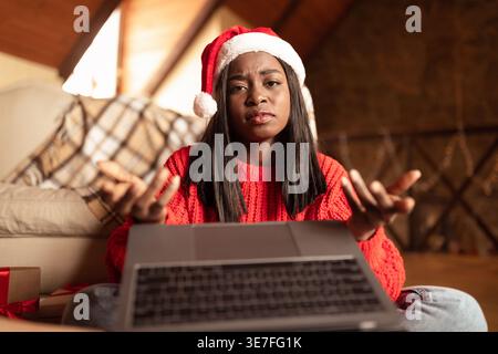 Bouleversée adolescente noire portant un chapeau de Père Noël faisant des gestes des mains tout en ayant un appel vidéo à l'aide d'un ordinateur portable assis sur le sol dans le salon à la maison Banque D'Images