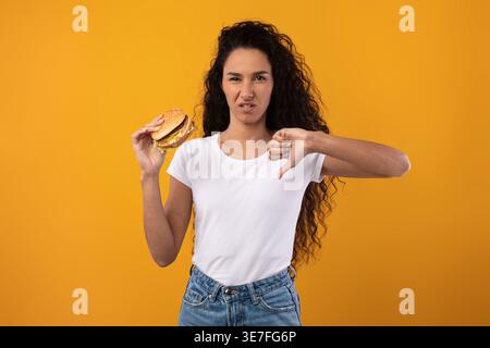 Femme du moyen-Orient avec les cheveux bouclés tenant un hamburger en faisant des gestes avec les pouces vers le bas tout en se tenant debout sur fond jaune dans le studio Banque D'Images