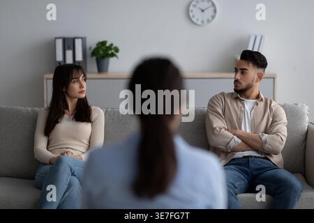 Couple moyen-oriental se regardant tout en étant assis sur le canapé devant la thérapeute féminine en clinique Banque D'Images