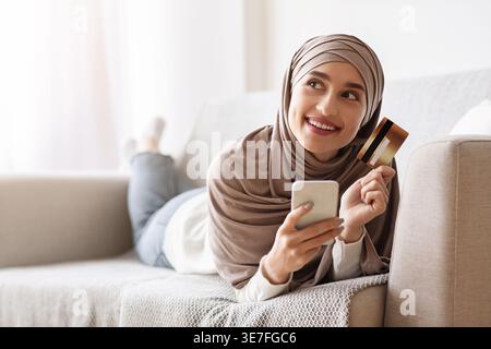 Femme musulmane arabe en hijab tenant un smartphone et une carte de crédit regardant de côté les achats en ligne allongée sur un canapé à la maison Banque D'Images