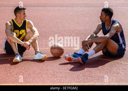 Des athlètes noirs et mixtes bavardant au repos et assis sur le sol sur un terrain de basket-ball par une journée ensoleillée Banque D'Images