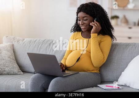 Femme enceinte noire avec des cheveux bouclés portant chemise jaune à manches longues à l'aide d'un casque ayant videocall sur ordinateur portable tout en étant assis sur le canapé dans le roo vivant... Banque D'Images