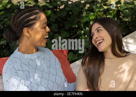 Diverses amies féminines assises sur un banc rembourré extérieur dans le jardin, discutant devant un mur de lierre Banque D'Images