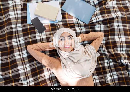 Étudiante musulmane indienne souriante portant le hijab regardant loin penchée la tête sur les deux mains tout en étant allongée sur une couverture à carreaux avec des livres et un smartphone... Banque D'Images