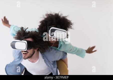 Couple noir et caucasien portant des casques VR ayant un plaisir piggyback équitation et expérimentant la réalité virtuelle sur fond blanc en studio Banque D'Images